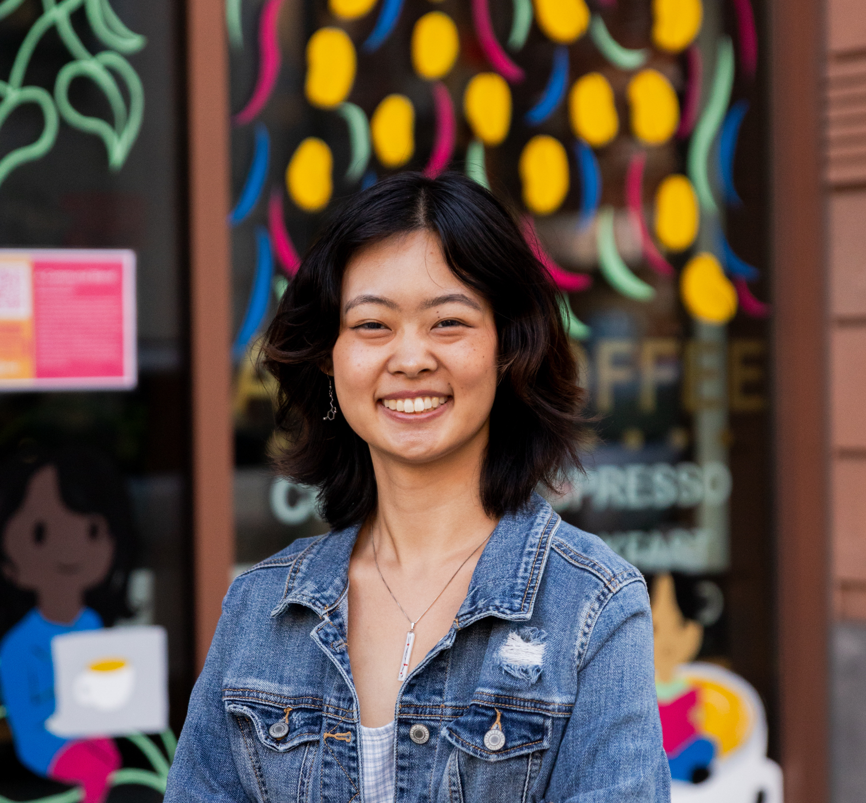 Headshot of Jennifer Duan. She is standing in front of the vibrantly decorated windows of a coffee shop.