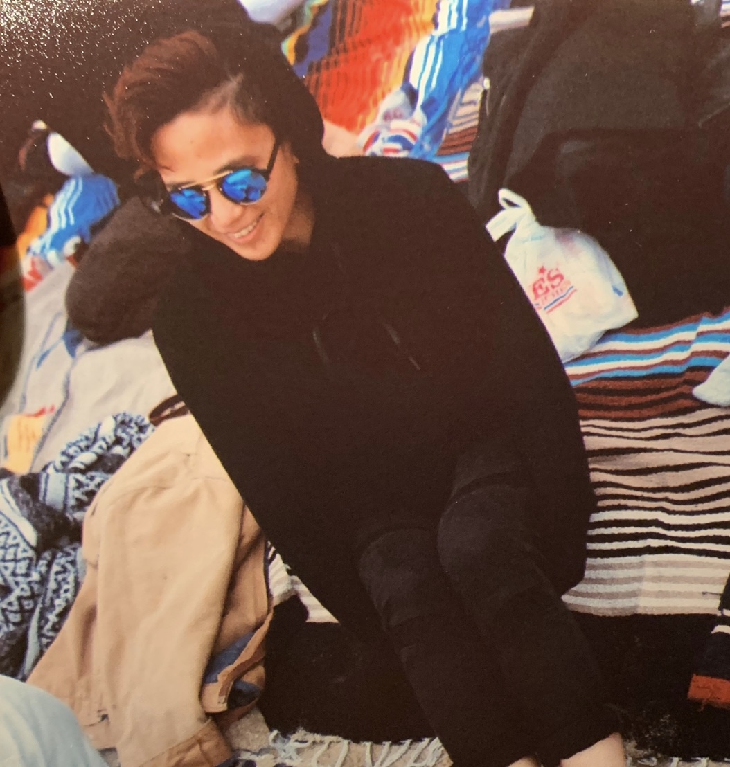 Close-up photo of Mikaela (Kelly) Carino. She is wearing blue-tinted sunglasses and sitting on a striped blanket.